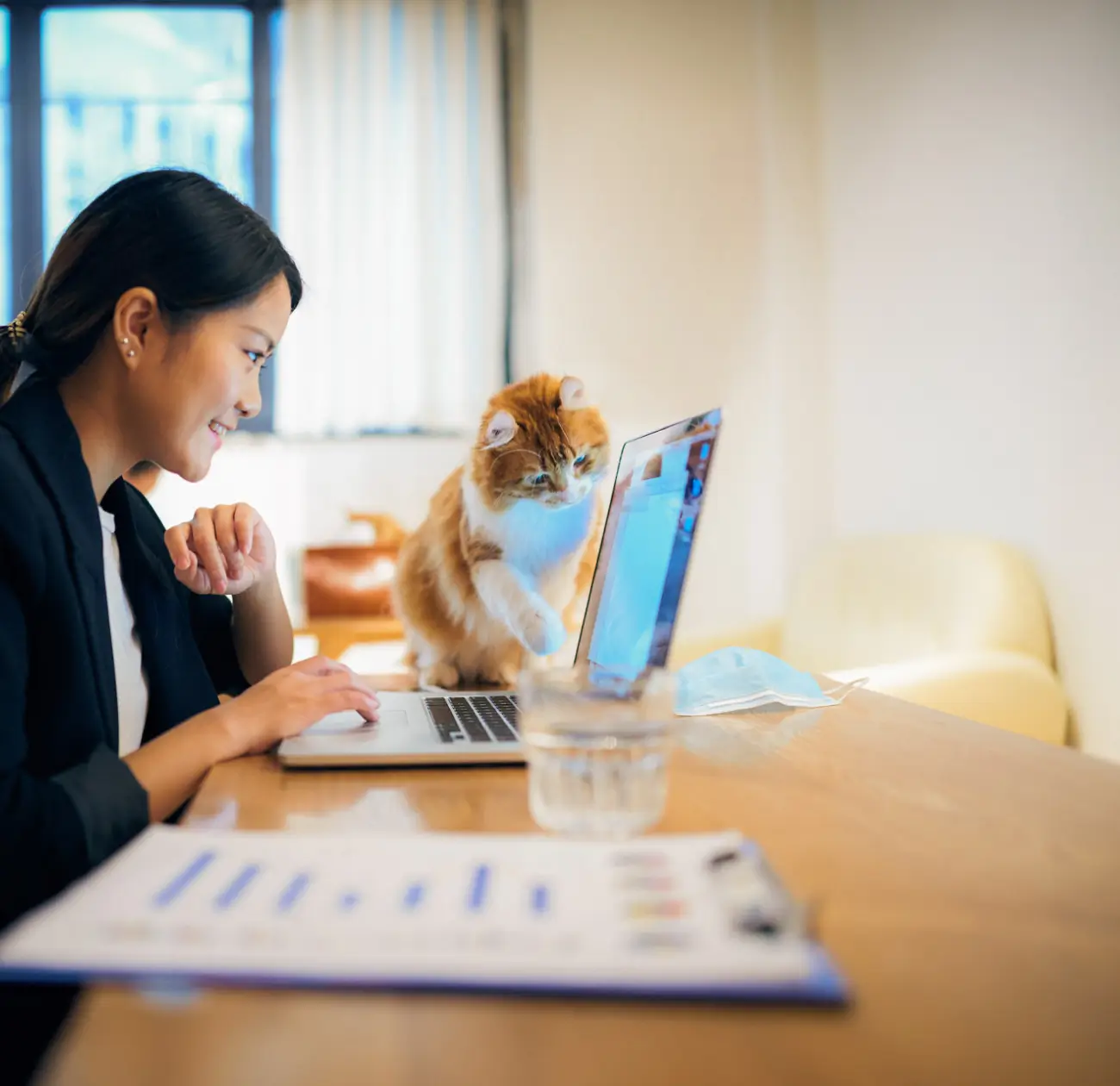 A happy employee works at a laptop in a bright office setting. Her furry cat sits next to her on the table, looking at the screen.