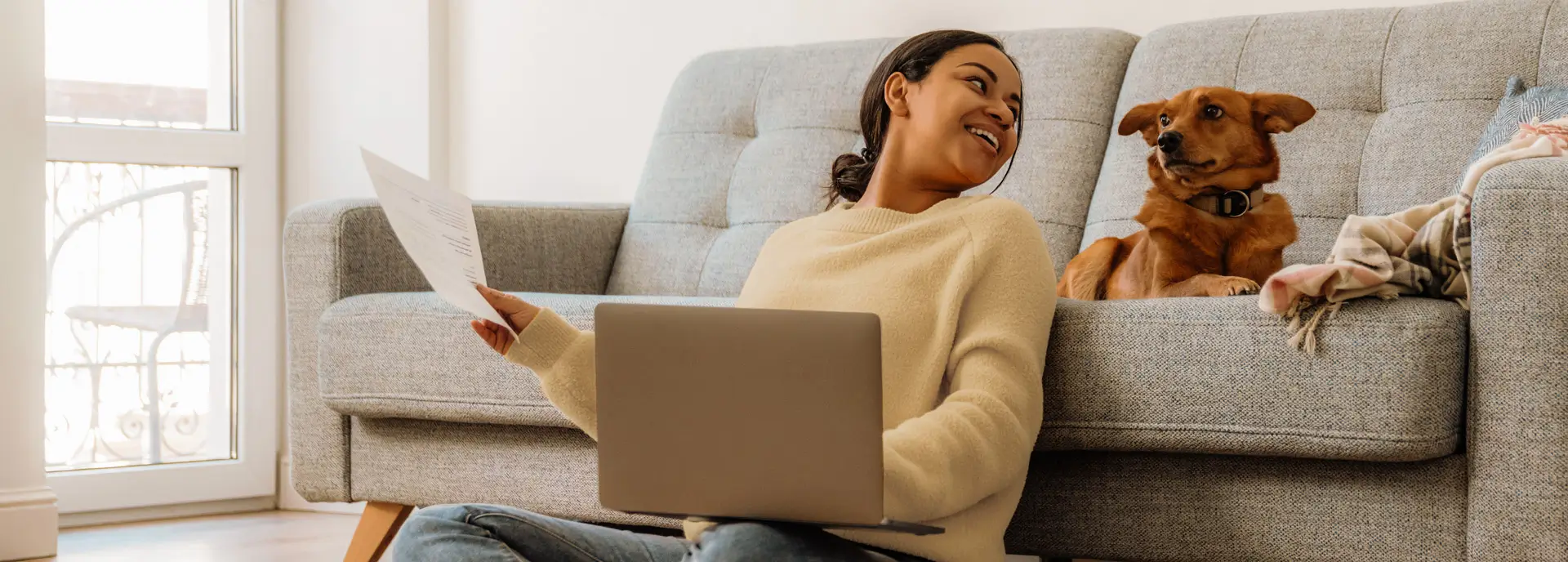 Woman sitting with her laptop on the floor, looking back at her dog relaxing on the couch