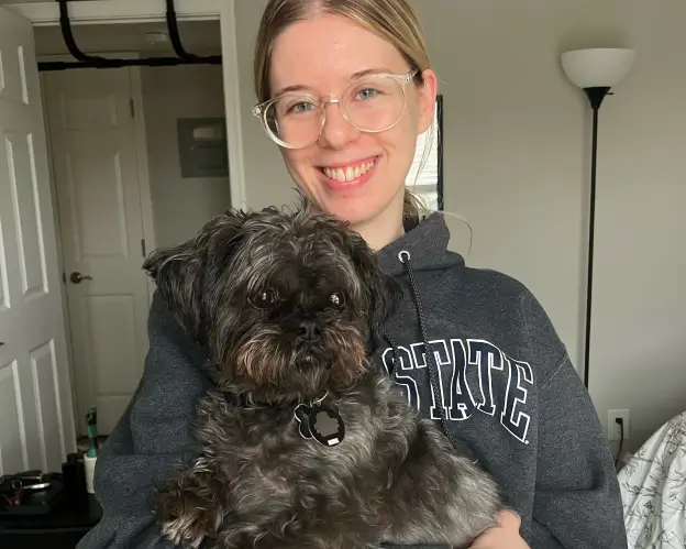 A young woman with glasses smiles brightly while holding a small black dog.